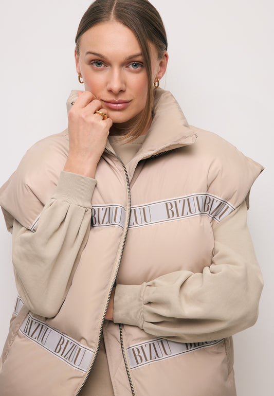 Zbliżenie na modelkę z ręką przy twarzy obraną w beżową kamizelkę puchową z logowaną taśmą AYASE.