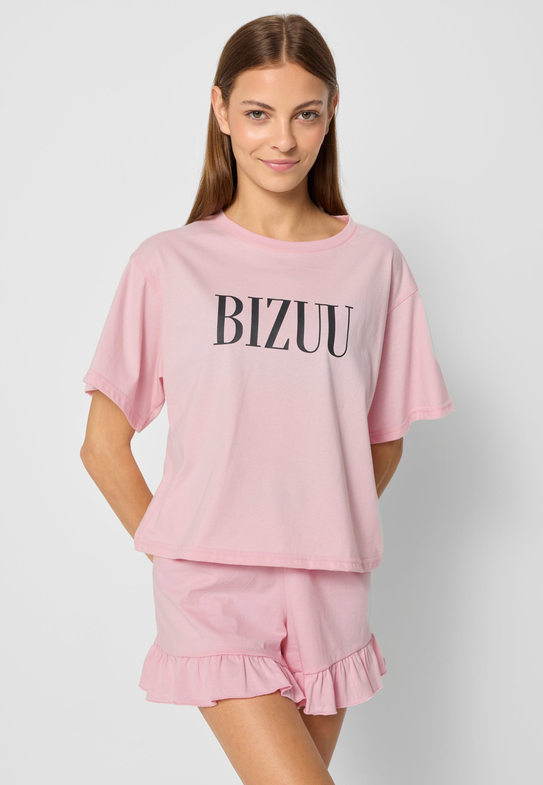 Modelka na tle białej ściany z rękoma z tyłu ubrana w różową piżamę z czarnym logo BIZUU.