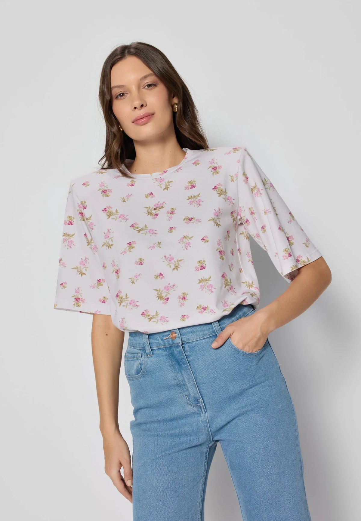 Niebieskie dżinsy noszone z białym t-shirtem w kwiaty, osoba stoi na neutralnym tle z ręką w kieszeni.