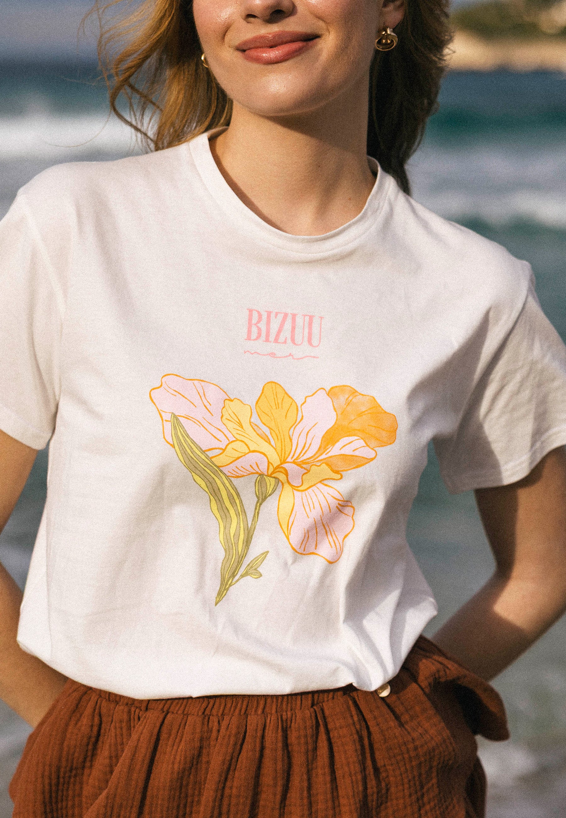 Zbliżenie na biały t-shirt TISAL z nadrukiem żółto-różowego kwiatu i różowym napisem BIZUU, noszony do brązowych szortów OSTER.