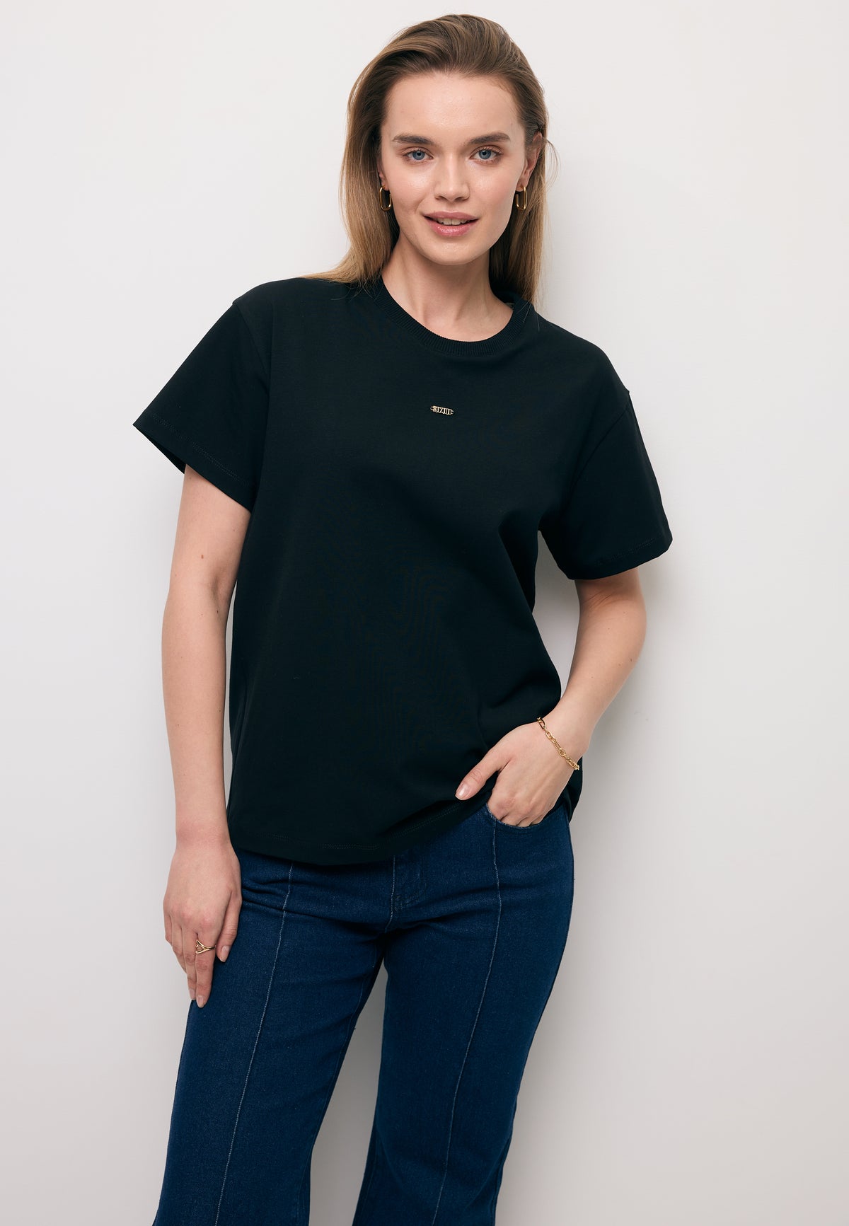 Modelka na tle białej ściany z jedną ręką na biodrze. Ubrana w czarny T-shirt TISHAL i koronkową spódniczkę FLACE.